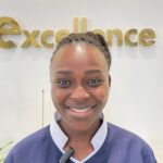 Portrait of a smiling woman in a navy uniform, standing in front of gold 'excellence' lettering on the wall.