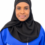 Portrait of a woman wearing a black hijab and blue blouse, smiling at the camera with an 'Excellence Center' logo on the shirt.