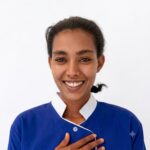 Smiling woman in blue scrubs with a white collar, hand over her heart against a plain white background.