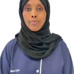 Portrait of a woman wearing a dark green hijab and a blue uniform, facing the camera with a neutral expression.